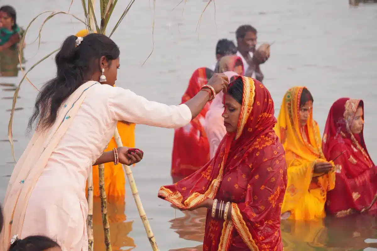 छठ पूजा 2026: व्रत कथा, चारों दिन की पूजा विधि, सामग्री सूची और ठेकुआ घाट पर पानी में खड़ी व्रतियाँ, एक लड़की व्रती को सिंदूर लगाती हुई